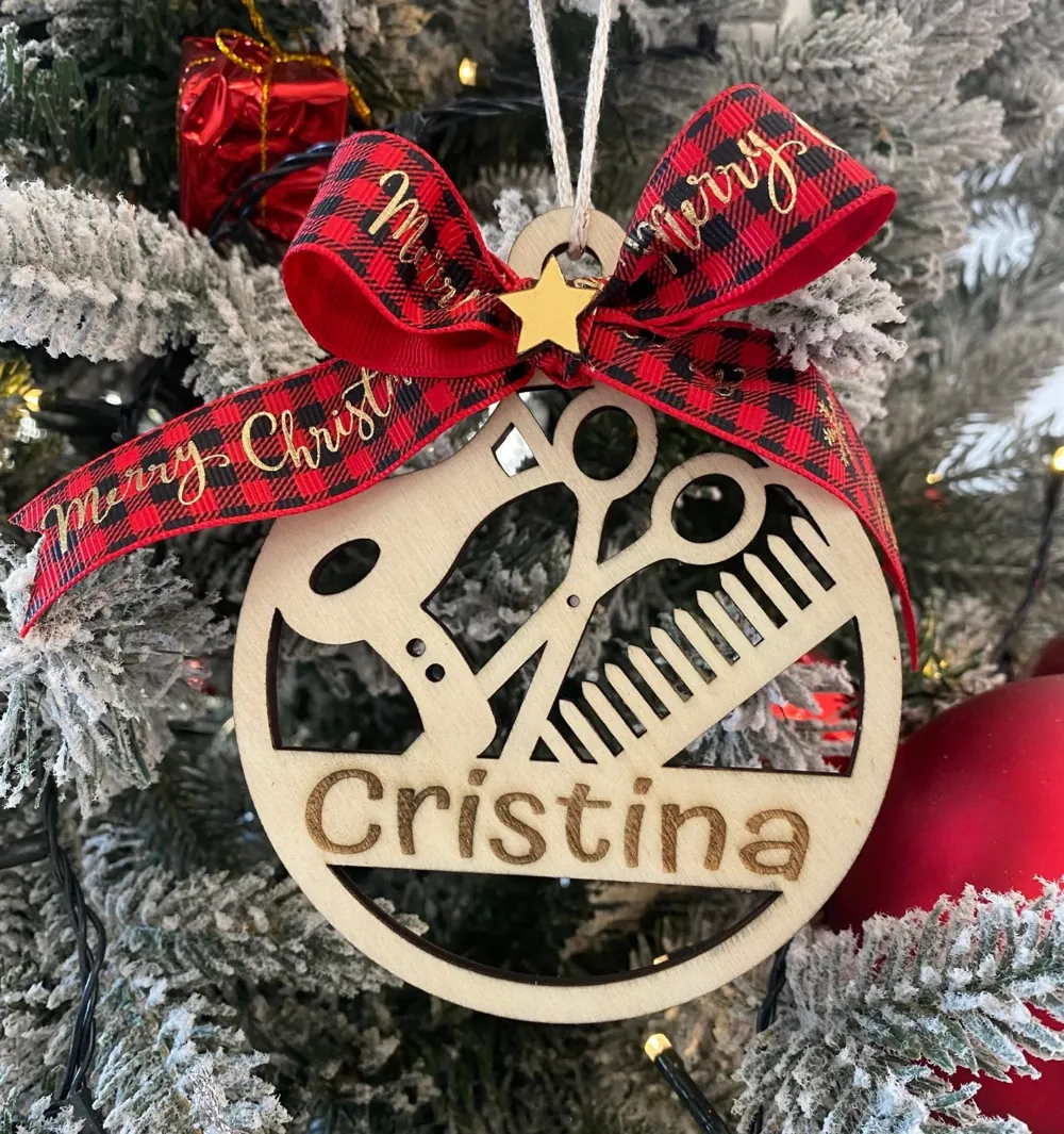 Pallina di Natale in legno con forbici e pettine traforati e il nome Cristina, sotto un fiocco rosso